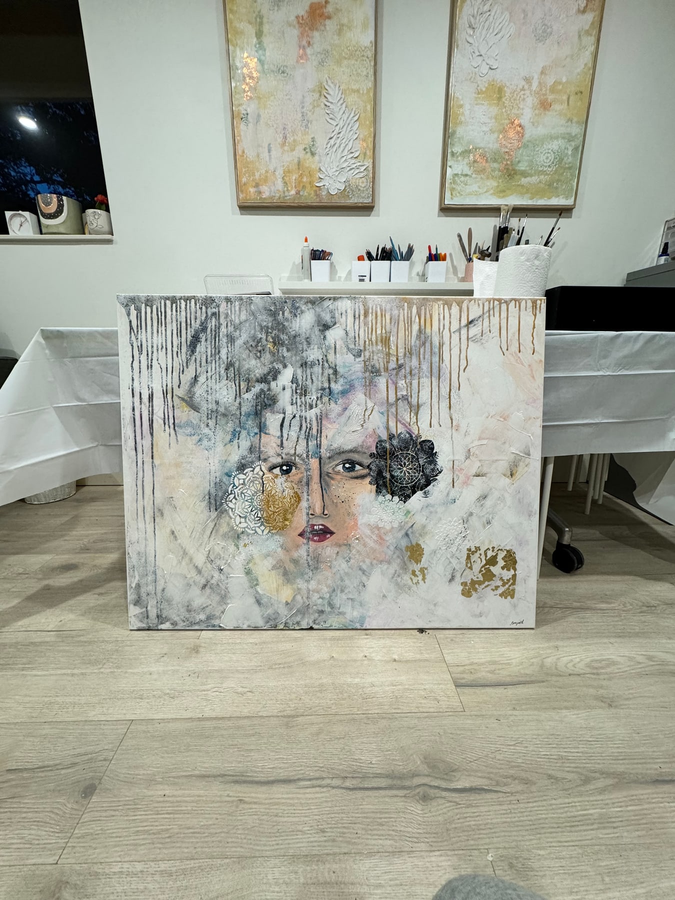 A large textured portrait canvas leaning against a bench in the studio, with framed work on the wall behind.